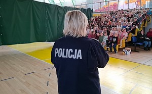 Policjantka przemawia do dzieci siedzących na ławkach w hali sportowej.
