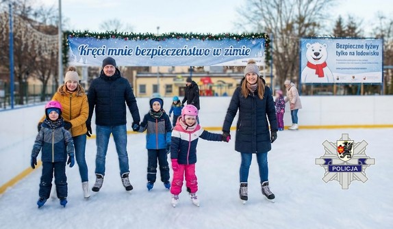 Rodzina z dziećmi na bezpiecznym lodowisku. W tle bilbordy z napisami: Kręci mnie bezpieczeństwo w zimie oraz Bezpieczne łyżwy tylko na lodowisku. W dolnym prawym rogu logo policyjne z herbem powiatu starogardzkiego.