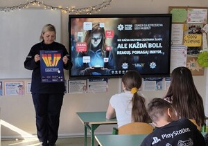 Policjantka w klasie szkolnej stoi i trzyma plakat dotyczący hejtu.