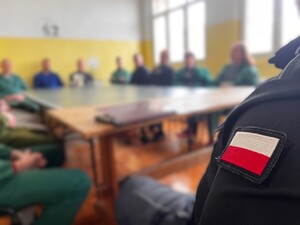 Biało-czerwona naszywka na lewym rękawie mundury Służby Więziennej, a w tle rozmazane wizerunki osób.