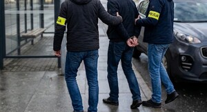 Policjanci po cywilnemu prowadzą mężczyznę, który ma założone kajdanki. W tle przystanek i nieoznakowany pojazd służbowy Policji.
