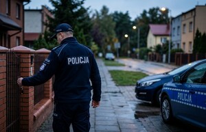 Policjant stoi przy furtce prowadzącej na prywatną posesję.