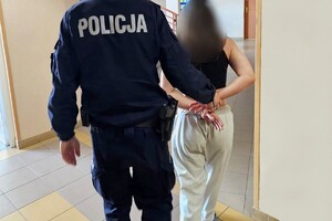Policjant prowadzi zatrzymaną kobietę. Kobieta ma założone kajdanki na rękach trzymanych z tyłu.