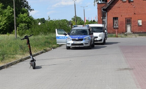 Na pierwszym planie stoi hulajnoga elektryczna. W tle radiowóz policyjny.