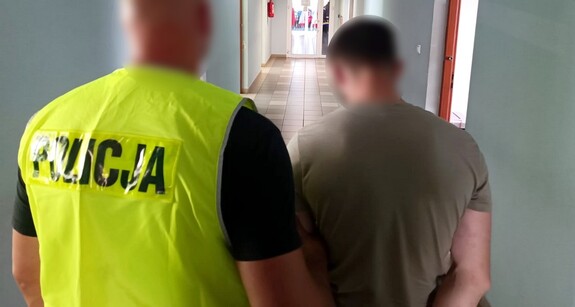 Policjant prowadzi zatrzymanego mężczyznę. W tle widoczny korytarz komendy.