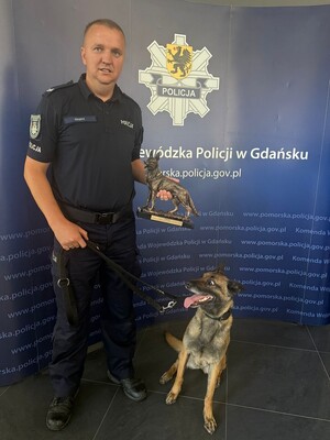 Przewodnik z psem służbowym. Policjant trzyma statuetkę, którą otrzymał za zajęcie drugiego miejsca.