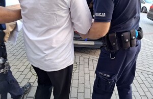 Policjanci prowadzą zatrzymanego mężczyznę w kierunku radiowozu.