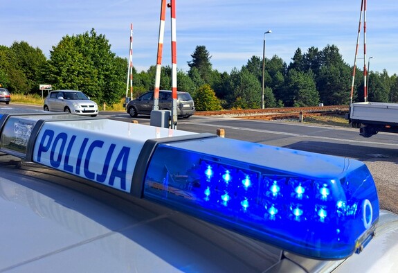 Kogut na dachu policyjnego radiowozu. Niebieski napis na białym tle o treści Policja. W tle widoczny przejazd kolejowy.