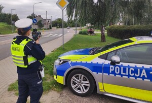 Policjantka z radarem. Po prawej stronie oznakowany radiowóz.